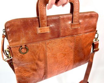 May include: A tan leather briefcase featuring a textured snakeskin pattern on the lower panels, two sturdy top handles, a top zipper closure, a brass-toned metal ring attachment on the front, and detachable side clips for an adjustable shoulder strap.