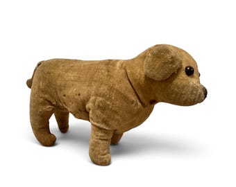 Puede incluir: Un juguete de perro de peluche de color tostado hecho de tela con una textura desgastada. El animal de peluche presenta una cabeza redondeada, orejas caídas, un pequeño hocico oscuro y dos ojos redondos de botón. Se sostiene sobre cuatro patas.