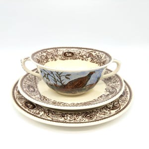 May include: A cream-coloured soup bowl with two handles, a saucer, and a plate. The bowl features a painted scene of a pheasant in a landscape. The set has a brown decorative border.
