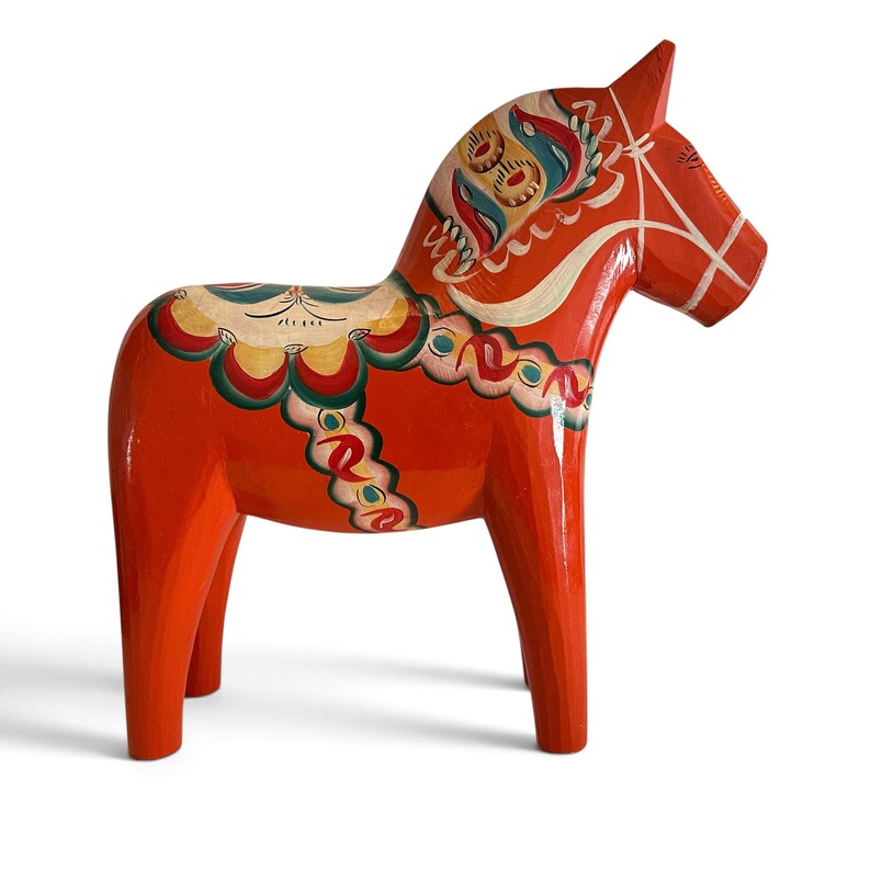 Puede incluir: Una figura de caballo Dala de madera de color naranja brillante con patrones decorativos pintados a mano en blanco, amarillo, verde y azul. El caballo presenta un dise&ntilde;o de silla de montar estilizado en el lomo y una brida pintada en la cabeza, con un acabado liso y brillante.