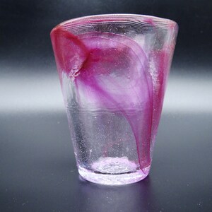 Puede incluir: Un vaso de vidrio transparente con forma acampanada, con vetas arremolinadas de color magenta y p&uacute;rpura. El borde tiene un dise&ntilde;o texturizado y acanalado. La base del vaso tiene un aspecto texturizado y esmerilado. El vaso est&aacute; sobre un fondo oscuro.