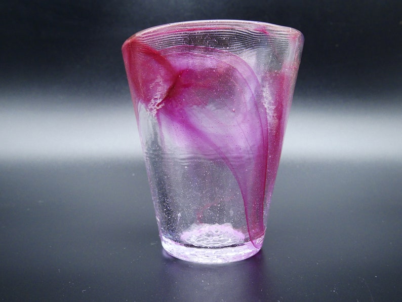 Puede incluir: Un vaso de vidrio transparente con forma acampanada, con vetas arremolinadas de color magenta y p&uacute;rpura. El borde tiene un dise&ntilde;o texturizado y acanalado. La base del vaso tiene un aspecto texturizado y esmerilado. El vaso est&aacute; sobre un fondo oscuro.