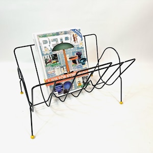 May include: Black metal magazine rack with a geometric design, holding a magazine with colorful images. The rack has yellow feet and a mid-century modern aesthetic.