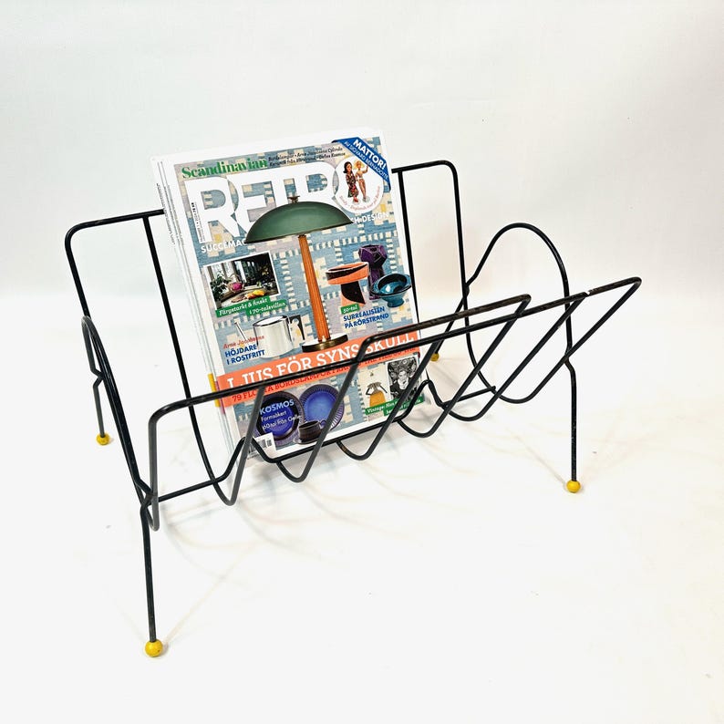 May include: Black metal magazine rack with a geometric design, holding a magazine with colorful images. The rack has yellow feet and a mid-century modern aesthetic.