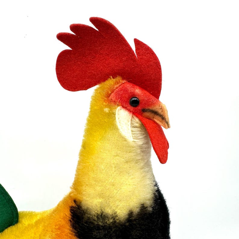 May include: A plush rooster toy with a vibrant red comb and wattle, yellow body, and black accents. The rooster has a detailed beak and a green tail feather. The toy is set against a white background.