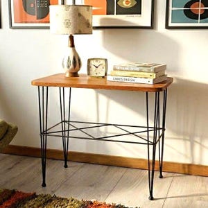 Könnte beinhalten: Ein Beistelltisch im Mid-Century-Stil mit rechteckiger Holzplatte und schwarzen Haarnadelbeinen. Eine Lampe, eine Uhr und Bücher stehen auf dem Tisch. Der Tisch hat eine untere Ablage aus schwarzen Metallstäben. Der Tisch ist ca. 76 cm lang.