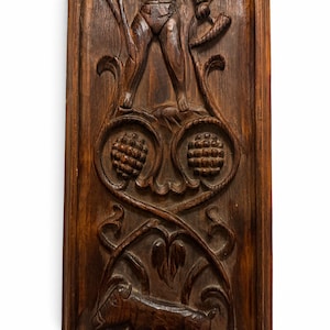 May include: A rectangular dark brown wooden wall panel featuring intricate relief carvings. The design includes a central figure standing above two circular motifs resembling pinecones, with a goat figure at the bottom base.