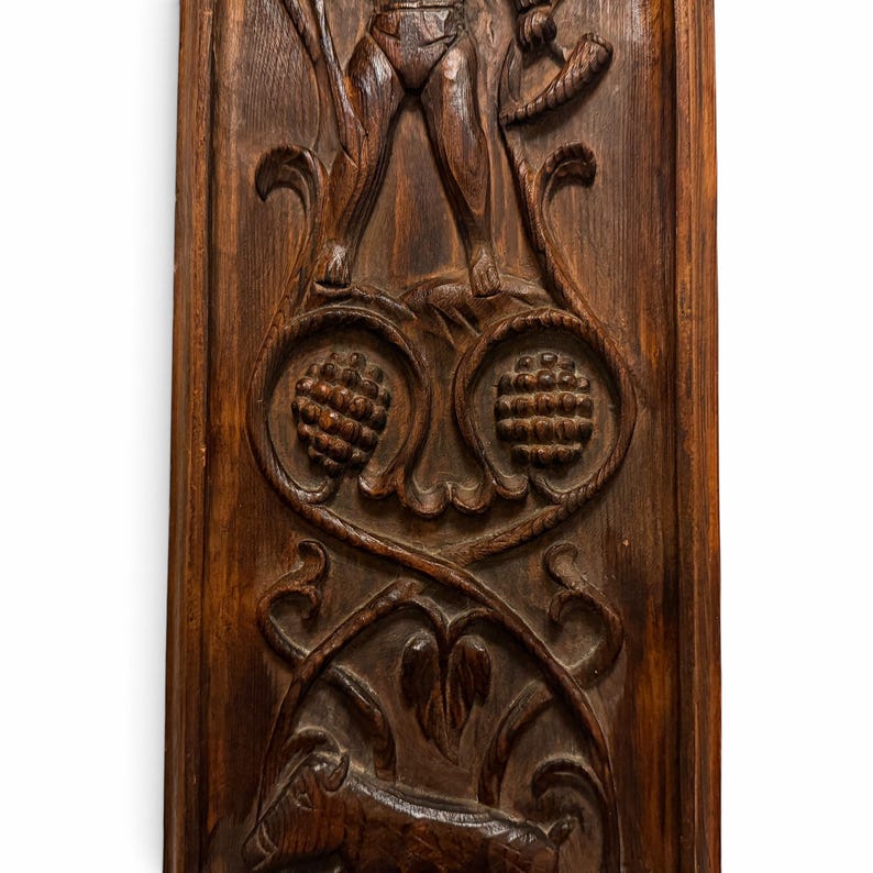 May include: A rectangular dark brown wooden wall panel featuring intricate relief carvings. The design includes a central figure standing above two circular motifs resembling pinecones, with a goat figure at the bottom base.