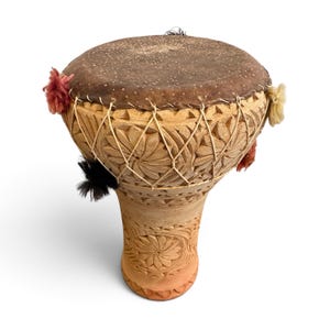May include: A goblet-shaped drum made of light brown carved clay with a dark brown animal skin head. The body features intricate floral patterns and is secured with white cord lacing. Small red, black, and cream-coloured yarn tassels are attached.