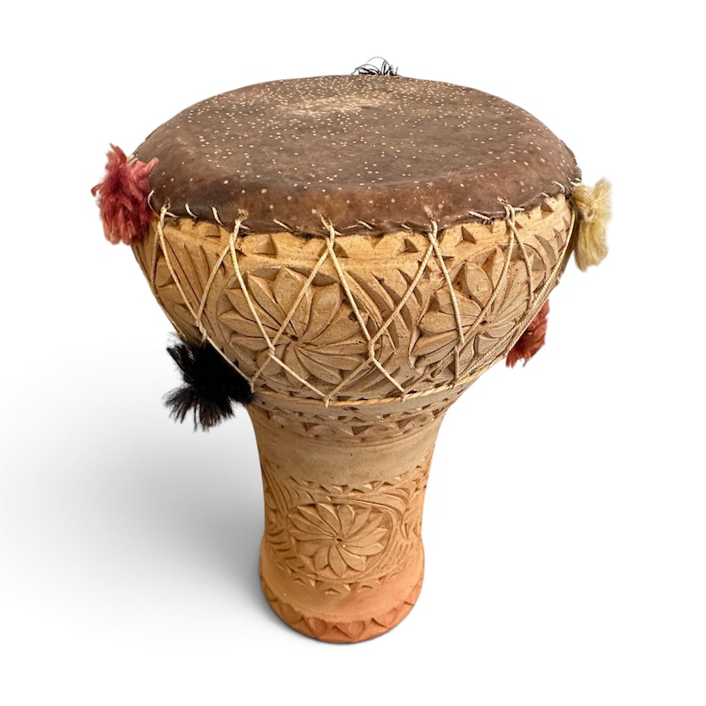 May include: A goblet-shaped drum made of light brown carved clay with a dark brown animal skin head. The body features intricate floral patterns and is secured with white cord lacing. Small red, black, and cream-coloured yarn tassels are attached.