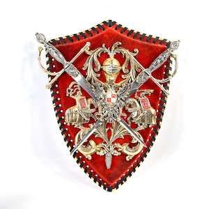 Könnte beinhalten: Ein schildförmiges Wandobjekt mit rotem Samthintergrund und einem schwarz geschnürten Lederrand. In der Mitte befindet sich ein goldfarbenes Metallemblem mit Helm, Doppeladler und zwei berittenen Rittern, überlagert von zwei gekreuzten silberfarbenen Schwertern.