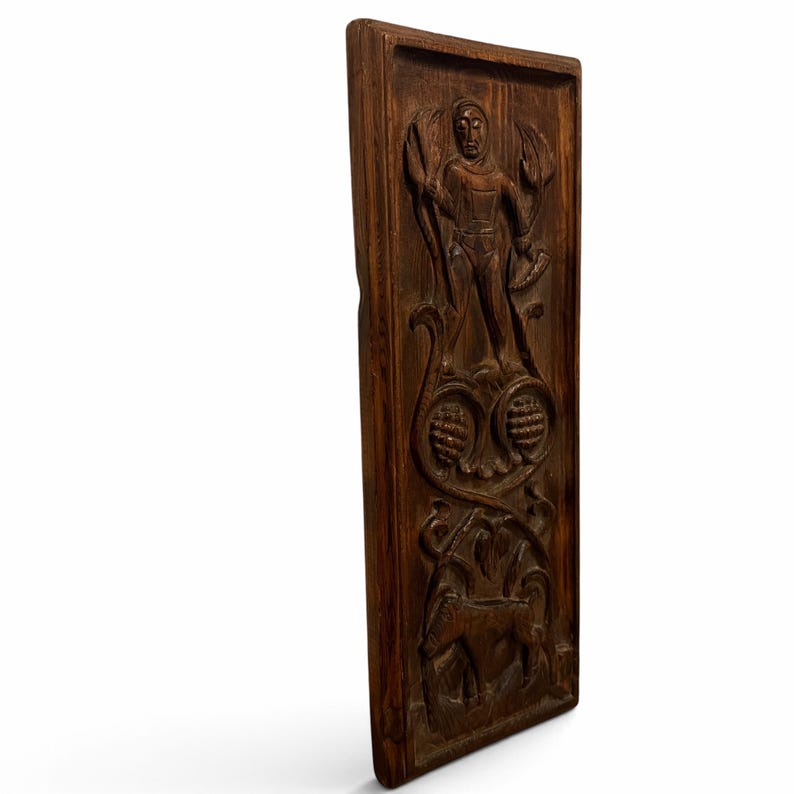 May include: A rectangular dark brown wooden wall plaque featuring a hand-carved relief design. The carving depicts a standing figure holding tools, flanked by stylized foliage, with two circular pinecone-like motifs and a four-legged animal below.