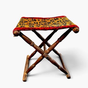 Op de afbeelding: Een opvouwbare kruk met een donkerbruin houten frame met gedraaide poten en een rechthoekige zitting bekleed met een stof met een zwart en goudkleurig bloemenpatroon, afgewerkt met een rode stoffen rand langs de zijkanten.
