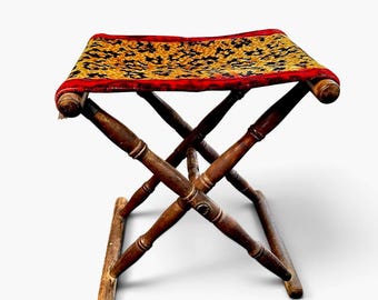 May include: A folding stool with a dark brown wooden frame featuring turned legs and a rectangular seat upholstered in a fabric with a black and gold floral pattern, finished with a red fabric border along the edges of the seat.
