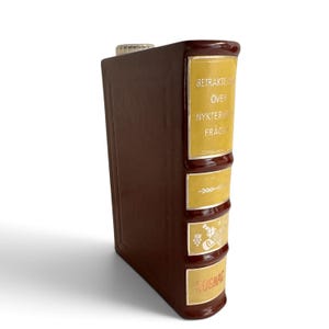 Puede incluir: Un decantador de cer&aacute;mica marr&oacute;n con forma de libro antiguo y lomo amarillo. El lomo presenta el texto plateado BETRAKTELSER &Ouml;VER NYKTERHETSFR&Aring;GAN, un gr&aacute;fico de una botella con uvas y la palabra COGNAC en la parte inferior con un tap&oacute;n claro.