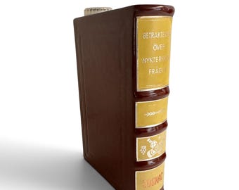 May include: A brown ceramic decanter shaped like a vintage book with a yellow spine. The spine features silver text reading BETRAKTELSER ÖVER NYKTERHETSFRÅGAN, a graphic of a bottle and grapes, and the word COGNAC at the bottom with a clear stopper.
