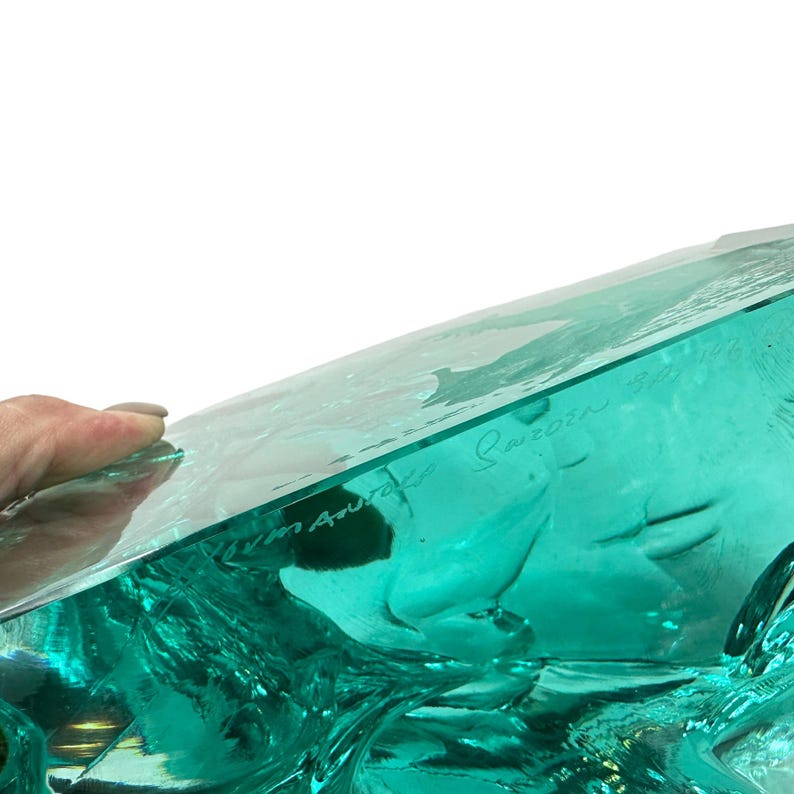 Peut inclure: Une sculpture en verre bleu sarcelle translucide et &eacute;pais, pr&eacute;sentant une forme organique irr&eacute;guli&egrave;re. La surface comporte une inscription grav&eacute;e "Mats Jonasson Sweden" sur le c&ocirc;t&eacute;, avec un bord sup&eacute;rieur plat et lisse.