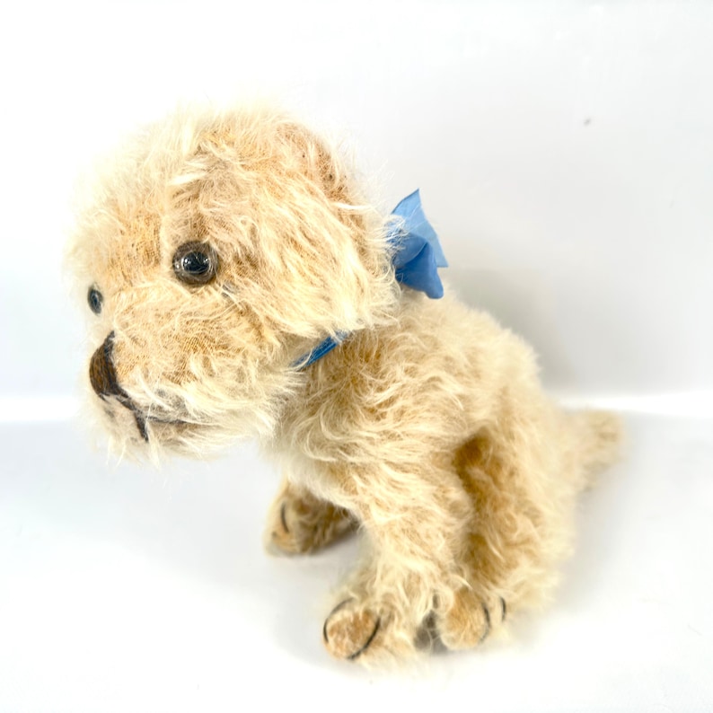 以下が含まれることがあります： 首に青いリボンを結んだ、タン色のぬいぐるみの子犬。 ぬいぐるみには黒い目と黒い鼻があります。 犬は後ろ足で座っており、前足は伸ばされています。