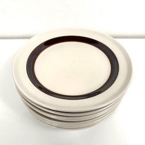 May include: A stack of six white ceramic dessert plates with a brown band and a white embossed pattern around the edge.