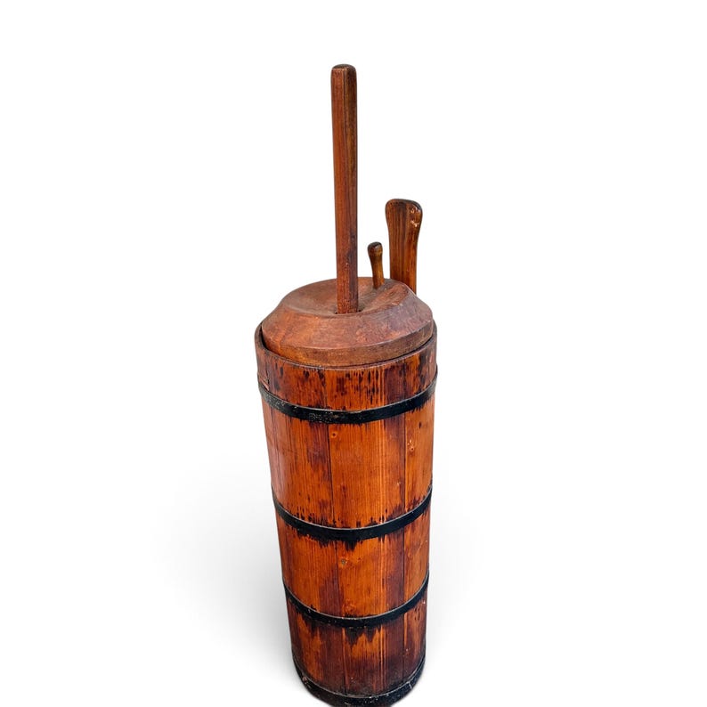 May include: A tall, cylindrical wooden butter churn featuring vertical staves held together by three black metal bands. It includes a circular wooden lid with a central vertical plunger handle and a smaller, separate wooden paddle tool attached.