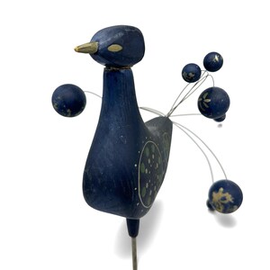 May include: A dark blue wooden bird figurine featuring a gold-painted beak and eyes. The bird has a long neck and a body decorated with green and gold circular patterns. Thin metal wires extend from the back, each topped with a small blue sphere.