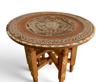 May include: A round wooden side table featuring intricate geometric inlay patterns on the tabletop surface. The table is supported by four legs connected by a cross-brace structure, with small inlaid floral motifs decorating the legs.