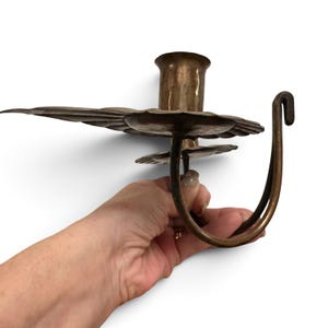 K&ouml;nnte beinhalten: Ein Kerzenhalter aus Metall mit geh&auml;mmertem Finish, bestehend aus einer zentralen zylindrischen Kerzenaufnahme, einer breiten, gewellten Tropfschale und einem geschwungenen Griff mit Hakenende, gefertigt in einem warmen, br&uuml;nierten Bronzeton.