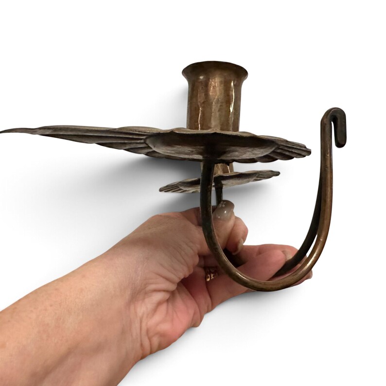 K&ouml;nnte beinhalten: Ein Kerzenhalter aus Metall mit geh&auml;mmertem Finish, bestehend aus einer zentralen zylindrischen Kerzenaufnahme, einer breiten, gewellten Tropfschale und einem geschwungenen Griff mit Hakenende, gefertigt in einem warmen, br&uuml;nierten Bronzeton.