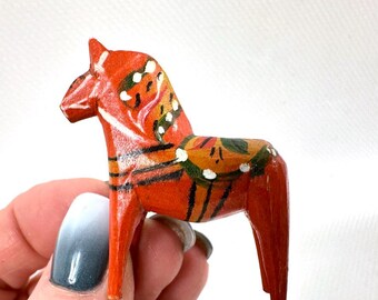 May include: A hand-painted, wooden Dala horse figurine. The horse is painted a vibrant red with decorative floral patterns in white, green, and yellow. The figurine is held in a hand against a white background.