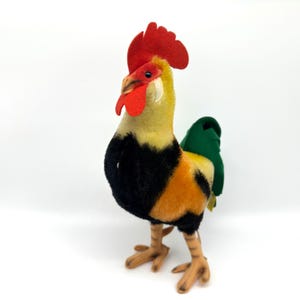 May include: A plush rooster toy with a red comb and wattle, yellow and black body, and green tail feathers. The rooster has tan legs and feet, and is standing on a white surface.