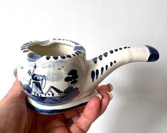 May include: A ceramic pipe-shaped ashtray featuring a hand-painted blue and white landscape design. The bowl displays a windmill and house scene with a crackled glaze finish. The long stem has blue decorative markings along the top surface.