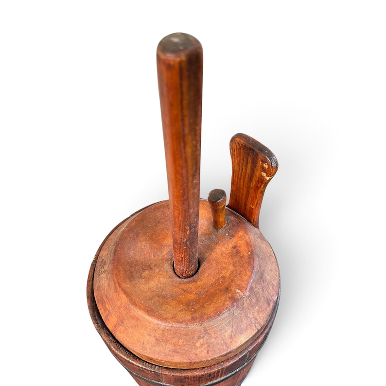 May include: A vintage wooden butter churn featuring a cylindrical body with a dark brown finish, a circular lid, and a central vertical plunger handle. The top assembly includes a small secondary handle and a curved wooden side piece.