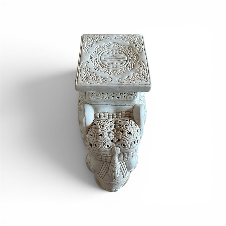 Może przedstawiać: Jasnoszary ceramiczny stołek ogrodowy w kształcie słonia. G&oacute;rna powierzchnia posiada kwadratową platformę ze skomplikowanym okrągłym wzorem reliefowym. Głowa słonia jest ozdobiona szczeg&oacute;łowymi, ażurowymi rzeźbieniami i motywami kwiatowymi.