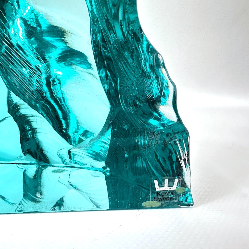 May include: A teal-colored art glass sculpture with a textured, organic form. The glass is translucent, allowing light to pass through, and features the text "Kosta Sweden" on a small label.
