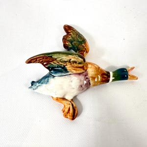 May include: A ceramic mallard duck figurine in flight, featuring a glossy finish with a dark green head, a white neck ring, brown and tan wings with green accents, a white underbelly, and orange webbed feet with its beak open.