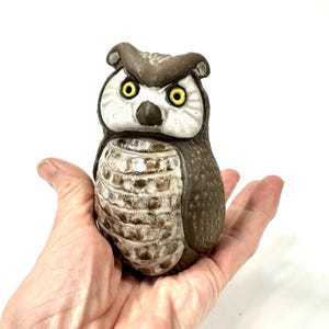 May include: A ceramic owl figurine in brown and white. The owl has large yellow eyes and a detailed feather pattern. The figurine is held in a hand against a white background. The owl is approximately 10 cm tall.