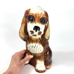Peut inclure: Figurine de chien en c&eacute;ramique, assis. Le chien a un pelage brun et blanc, de grands yeux expressifs et des oreilles tombantes. La figurine est peinte &agrave; la main avec une finition brillante, et le nez du chien est noir.