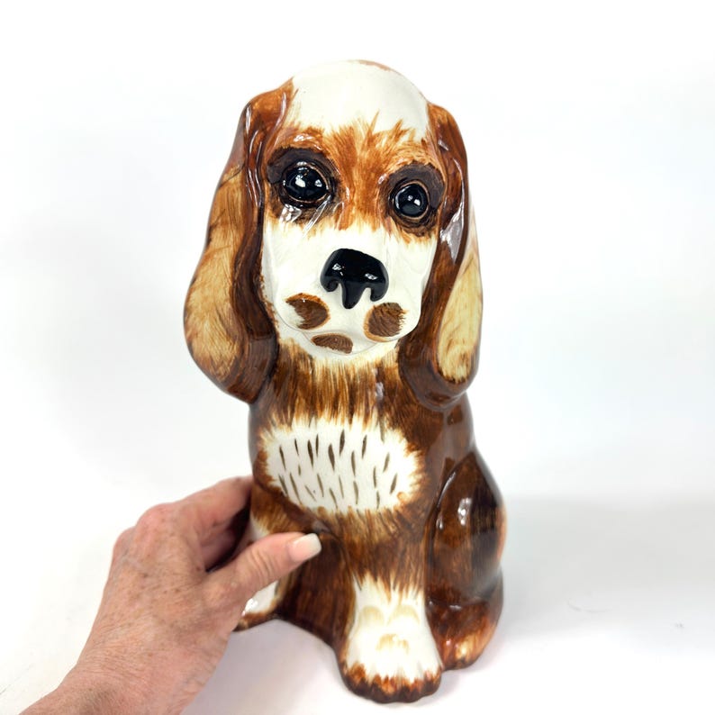 Peut inclure: Figurine de chien en c&eacute;ramique, assis. Le chien a un pelage brun et blanc, de grands yeux expressifs et des oreilles tombantes. La figurine est peinte &agrave; la main avec une finition brillante, et le nez du chien est noir.