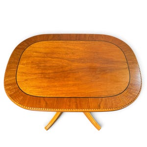 Op de afbeelding: Een ovale houten tafel met een glad, lichtbruin oppervlak met een duidelijk houtnerfpatroon. Het tafelblad is omlijst door een donkerdere houten rand met een kralendetail en wordt ondersteund door twee schuin naar buiten lopende houten poten.