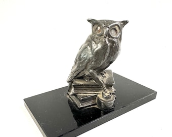 May include: A silver-toned metal owl figurine perched atop a stack of two books, with one talon resting on a small circular base. The entire sculpture is mounted on a rectangular, polished black base with a smooth, reflective surface finish.
