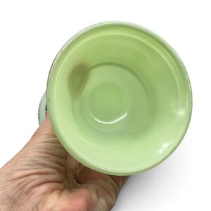 K&ouml;nnte beinhalten: Ein hellgr&uuml;ner Glasbecher mit glattem, abgerundetem Innenraum und leicht ausgestelltem Rand. Das Glas hat eine durchscheinende Beschaffenheit und zeigt eine subtile Variation in der Farbdichte sowie einen kleinen, dunkleren kreisf&ouml;rmigen Fleck.