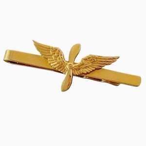 Könnte beinhalten: Eine goldfarbene Krawattenklammer aus Metall mit einem zentralen Emblem, das einen Flugzeugpropeller mit zwei ausgebreiteten Flügeln zeigt. Die Krawattenklammer hat eine glatte, polierte Oberfläche und das Luftfahrtmotiv ist mittig platziert.