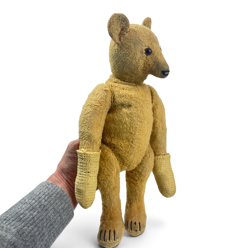 Pode incluir: Um urso de peluche dourado-bronzeado com corpo de tecido texturizado, detalhes de nariz e boca costurados em castanho escuro, olhos pequenos e escuros, e luvas de malha amarelo-claro nas patas, com marcas de garras costuradas nos p&eacute;s.