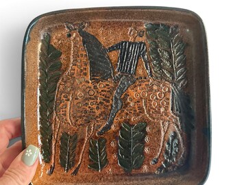 May include: A square ceramic dish with rounded corners featuring a brown glazed surface. The centre depicts a dark brown incised illustration of a person riding a horse, surrounded by stylised dark green leaf patterns and small star-like motifs.