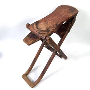 May include: A vintage folding wooden stool with a weathered brown finish. The seat is rectangular with a curved edge and a semi-circular wooden detail on top. The base consists of two intersecting wooden legs that form an X-frame structure.