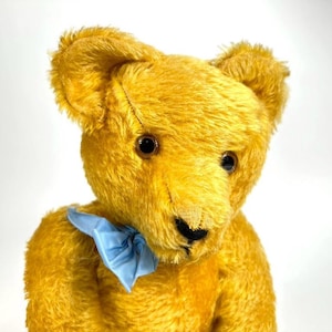 May include: A vintage golden-yellow teddy bear with dark brown eyes and a black stitched nose. The bear has a light blue bow tied around its neck. The plush toy is made of a soft, textured material.