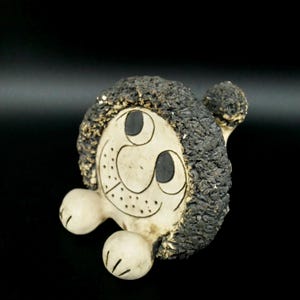 Può includere: Una statuetta in ceramica di una creatura sorridente con occhi neri e una criniera nera e pelosa. La creatura ha un corpo bianco e due piccole zampe bianche.
