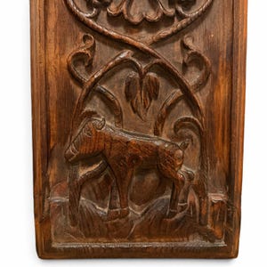 May include: A rectangular dark brown wooden panel featuring a relief carving of a four-legged animal standing in a grassy area. Above the animal, the wood is carved with swirling vine-like patterns and leaf motifs within a circular border.