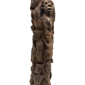 May include: A tall, dark brown wooden sculpture featuring a vertical arrangement of multiple human figures carved in relief. The figures are stacked and intertwined, displaying various facial expressions and postures, with a textured, matte finish.