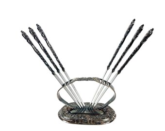 May include: A set of six silver-toned metal cocktail picks with ornate, twisted handles, arranged in a matching oval-shaped metal stand. The base and the handle of the stand feature intricate embossed floral patterns and a polished metallic finish.